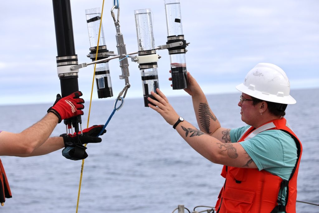 A postdoctoral researcher wearing a white hard hat and orange life vest prepares to deploy a scientific instrument from the deck of a research ship.