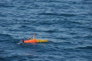 An orange-and-yellow-colored, torpedo-shaped, autonomous robot deployed in the ocean. The robot is at the ocean’s surface, with two orange fins and a black propeller sticking out of the water. Rippled blue ocean is visible in the background.