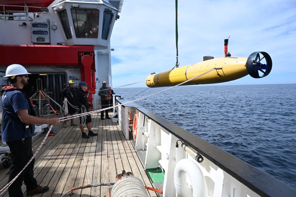 Four marine operations crew members deploy an autonomous robot over the side of a research ship. The crew are wearing hard hats and life vests and standing on the wooden deck of a research vessel on the left side of the frame. On the right is a cylindrical robot with a yellow plastic housing and black propellers suspended from a green nylon rope and a crane out of frame. The background is gray-blue ocean with cloudy blue sky on the horizon.