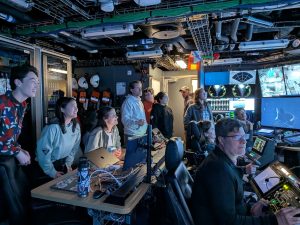 Scientists, submersible pilots, and ship’s crew gather in the dark submersible control room aboard a research ship, watching a live video feed of a deep-sea barreleye fish observed by an underwater robot.