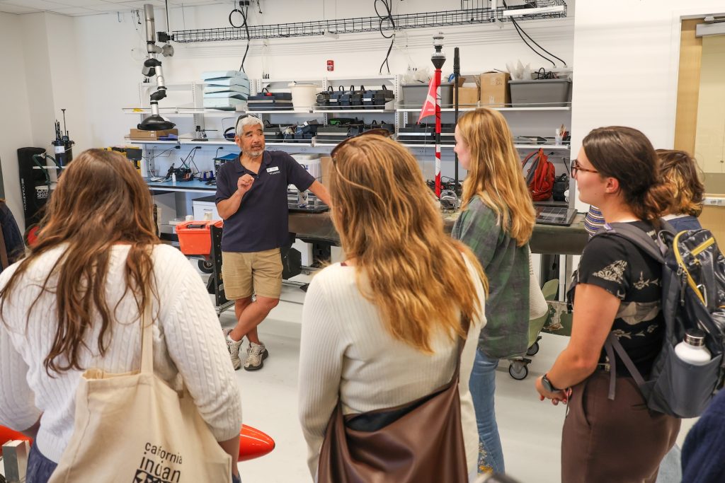 An MBARI researcher tours nine research interns and staff through an engineering lab. He is standing next to a dark-green autonomous robot, facing the interns who have their backs to the camera. Behind him are white walls lined with silver metal shelves and assorted engineering equipment.