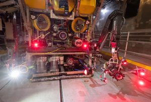 A deep-sea robot undergoes system checks prior to deployment. The robot has a yellow float at the top, black metal frame with two red lights in the middle, and silver metal frame at the base. On the left is a bright white light. On the right is a camera system in a black plastic housing shining five bright red lights. The background is the silver metal floor of a research vessel.