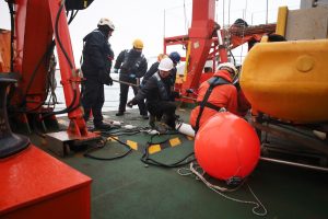Five marine operations crew members prepare a scientific instrument for deployment from a research ship. The crew are wearing hard hats and insulated jumpsuits. Some are standing, and some are kneeling on the ship deck while adjusting wires. In the foreground are a bright orange float and the green metal deck of the ship. In the background are various pieces of maritime equipment with gray-blue ocean, ice-covered mountains, and overcast gray sky on the horizon.