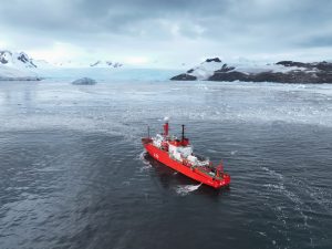 A polar research ship sails in waters around Antarctica. The ship has a bright reddish-orange hull and is moving away from the camera. In the foreground is grayish-blue ocean with several small drifting pieces of white ice. In the background are a large white glacier, mountains covered in white ice and snow, and overcast gray sky.
