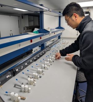 A researcher processes water samples in a lab on a research ship. The researcher has short black hair and is wearing glasses, a black jacket, and dark blue jeans. He is standing in front of a gray counter, adjusting several small clear plastic vials connected to a gray plastic cylinder by clear plastic tubing. In the background are a blue metal shelf and the white walls of the lab.