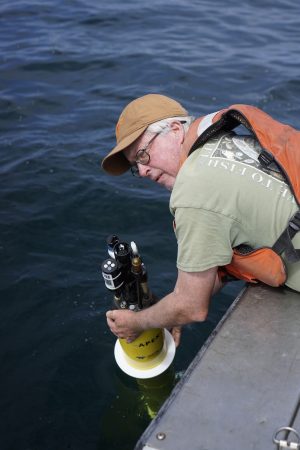 A researcher prepares to deploy a robotic float over the side of a small boat. The researcher is wearing a tan cap, green t-shirt, and an orange life vest. He is leaning over the silver metal side of a boat and holding the yellow plastic casing of a robotic float. In the background is still greenish-blue ocean.