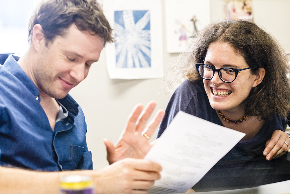 A musician and an educator seated at a table discuss an educational lesson plan in a brightly lit classroom.