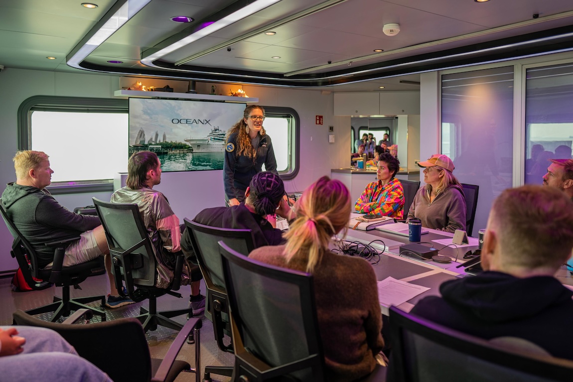 An educator leads an interactive workshop for a group of people in a conference room. The participants are seated around a table with their computers.