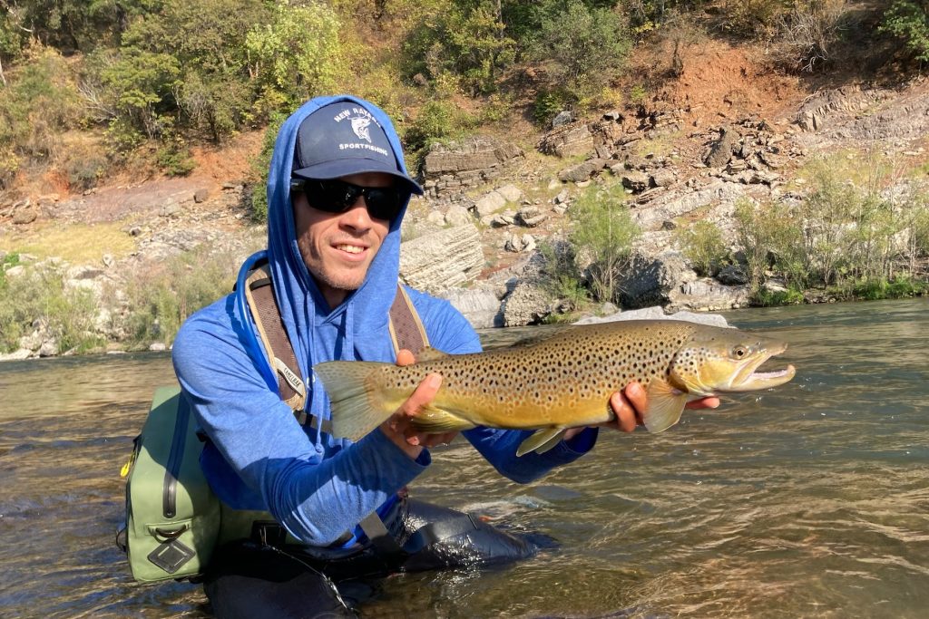 A scientist crouches in a shallow freshwater stream, posing for a photo while holding a trout in his hands. In the background is a reddish-brown dirt riverbank with scattered green trees.