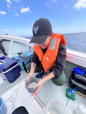 A scientist wearing an orange life vest crouches down on the gray deck of a small boat to examine a sample of seawater and plankton. She is surrounded by several bags of scientific equipment on the ship’s deck. In the background is blue ocean with cloudy blue sky on the horizon.