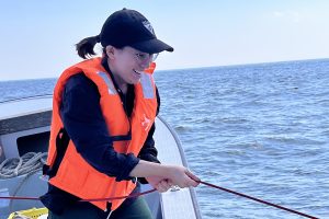 A scientist standing on a small boat wearing an orange life vest pulls a red rope to retrieve a sampling instrument. In the background is blue ocean with hazy blue sky on the horizon.