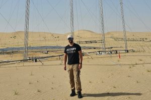An engineer wearing a cap and sunglasses stands in front of tall silver metal power infrastructure in the brown sandy desert.