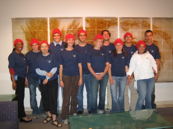 Twelve summer interns wearing blue t-shirts and red beanie caps pose for a photo in an office building. In the foreground are a glass table and a purple armchair. In the background is an art installation with dried brown kelp.
