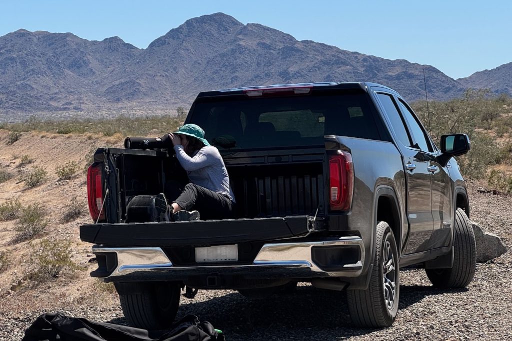 An engineer sits in the bed of a large black pickup truck and uses a telescope to observe a field experiment in the distance on a sunny day. The truck is parked on a gray gravel dirt road in a sandy desert, with mountains and blue sky in the background.