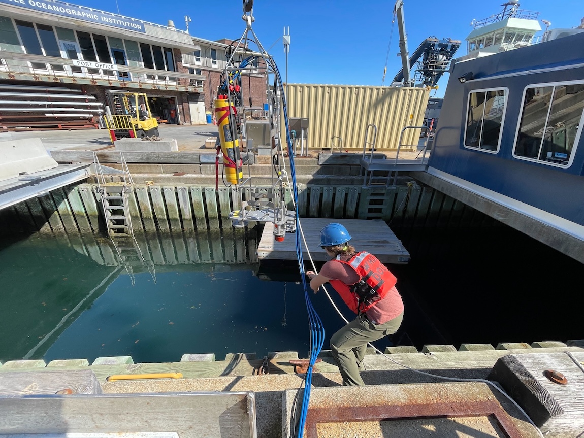 An engineer wearing a blue hard hat and orange life vest hoists a scientific instrument into position over a large pool of green water. The instrument has a yellow plastic housing and is attached to a silver metal frame. The engineer has her back to the camera and is holding a gray rope and bundle of blue cables connected to the instrument. In the background are office buildings, with clear blue sky in the distance.
