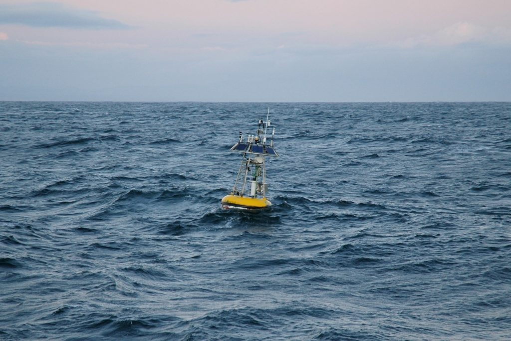 A yellow buoy with scientific instruments floats at the ocean’s surface in open blue water.