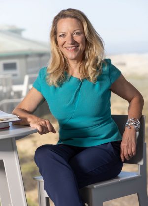 MBARI President and CEO Antje Boetius sits at a high coffee table. She has shoulder-length blonde hair and is wearing a bright turquoise shirt, navy blue pants, and a silver metal bracelet. She is sitting on a gray chair and leaning on a gray coffee table with two science textbooks. In the background are an MBARI research building, the beach, and blue sky.