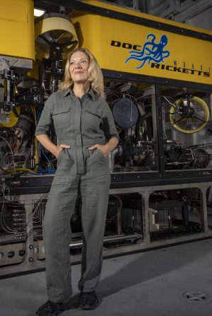 MBARI President and CEO Antje Boetius stands in front of a robotic submersible. She has shoulder-length blonde hair and is wearing an olive-green linen jumpsuit. The submersible has a yellow float with the name and logo reading Doc Ricketts above a silver metal frame with assorted electronics and scientific equipment.