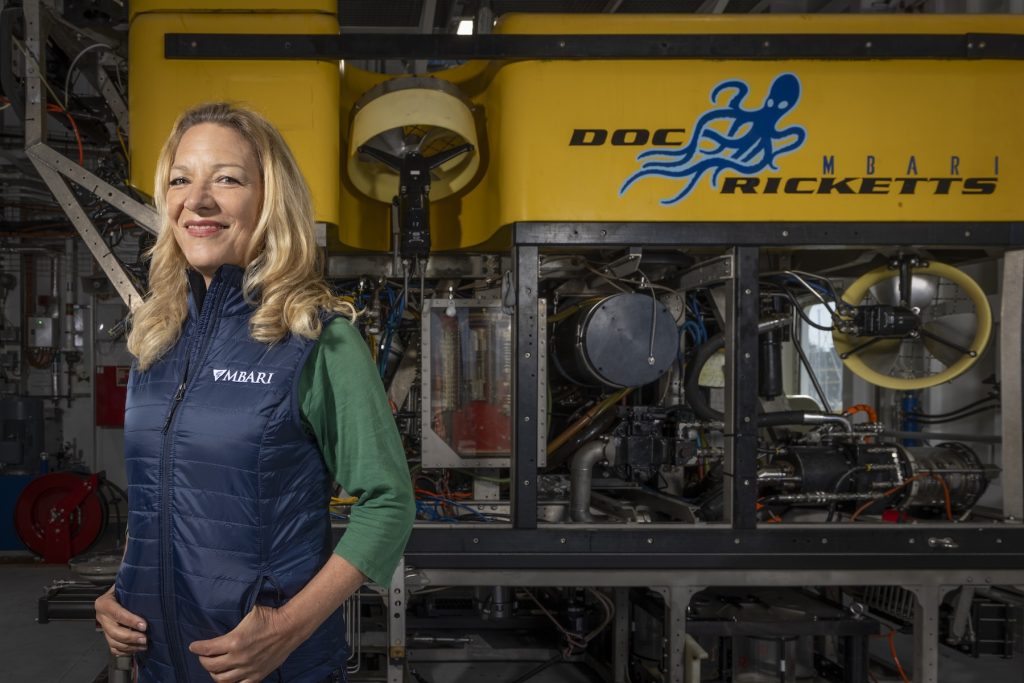 A scientist poses for a portrait in front of a robotic submersible inside a hangar aboard a research ship.
