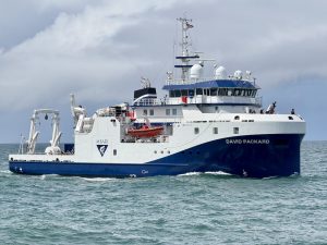An MBARI research vessel sails in greenish-blue water offshore of MBARI’s facilities. The research vessel is white with a blue hull printed with the MBARI logo and the vessel name DAVID PACKARD. On the deck are cranes and other oceanographic research equipment. In the background are gray clouds and pale-blue sky.