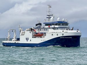 An MBARI research vessel sails in greenish-blue water offshore of MBARI’s facilities. The research vessel is white with a blue hull printed with the MBARI logo and the vessel name DAVID PACKARD. On the deck are cranes and other oceanographic research equipment. In the background are gray clouds and pale-blue sky.