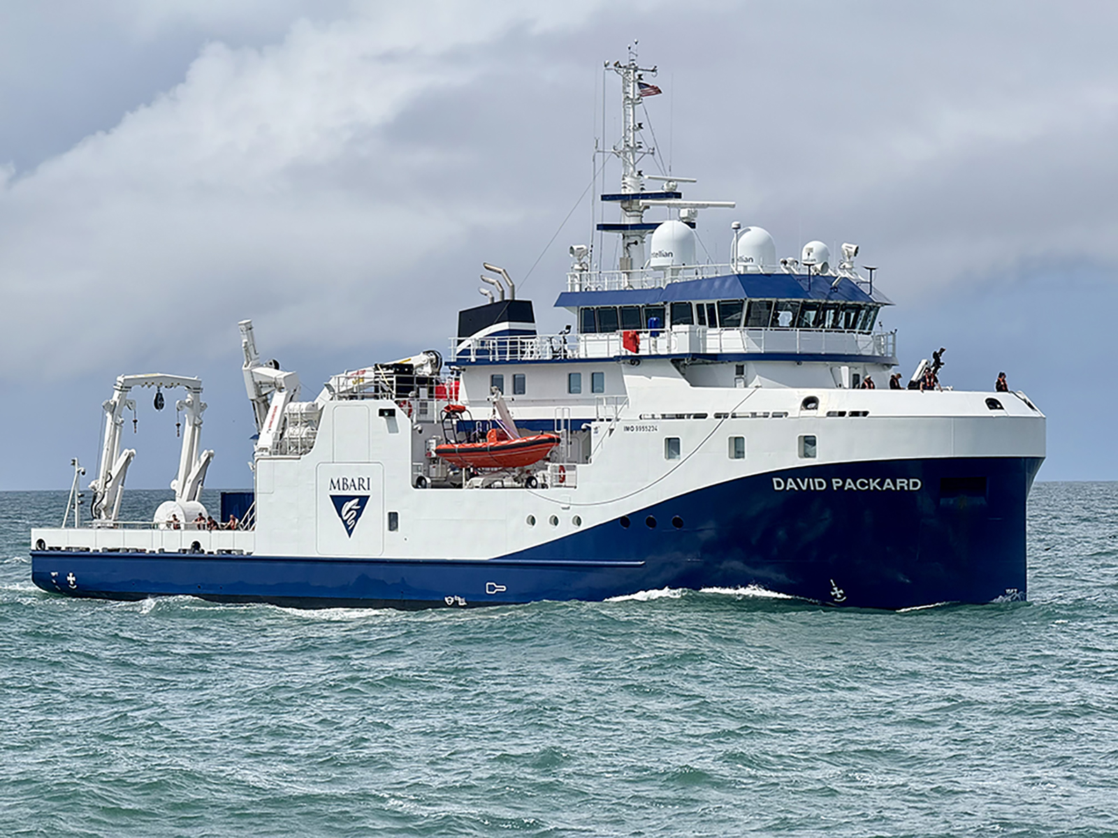 An MBARI research vessel sails in greenish-blue water offshore of MBARI’s facilities. The research vessel is white with a blue hull printed with the MBARI logo and the vessel name DAVID PACKARD. On the deck are cranes and other oceanographic research equipment. In the background are gray clouds and pale-blue sky.