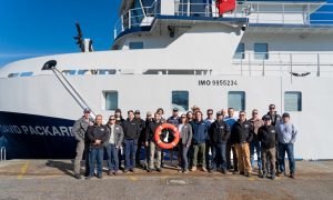 Twenty-three members of MBARI’s marine operations team stand in front of an MBARI research vessel. The group is standing on a rusty-brown cement dock, and two individuals in the center of the group are holding an orange flotation ring. The research vessel in the background is white with a blue hull, printed with the name DAVID PACKARD and a registration number. In the background is bright-blue sky.