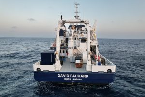 An MBARI research vessel sails in open grayish-blue water. The research vessel is white with a blue hull printed with the vessel name DAVID PACKARD and port MOSS LANDING. On the deck are cranes and other oceanographic research equipment. In the background is a hazy orange-and-blue sky.