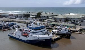 Two MBARI research vessels sit in port across the street from MBARI’s research facilities. A white research vessel with a blue hull is in the foreground, and a white research vessel with a black hull docked behind it. The research facilities include five large two-story gray buildings with several cars parked in front of each. In the foreground is the brown water of the harbor. In the background are greenish-blue ocean, white waves, light-blue sky, and gray clouds.