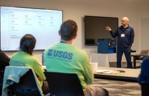 An engineer with a bald head and gray beard, wearing glasses, a blue pullover with the MBARI logo, and blue jeans, presents to workshop participants in a conference room. The engineer is looking at a screen displaying data. In the foreground are two workshop participants wearing bright green shirts with the USGS logo.