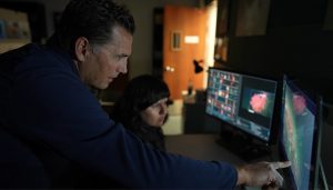 Two MBARI researchers look at two computer screens displaying labeled images used to train artificial intelligence to identify deep-sea animals. The researcher in the foreground has short brown hair, is wearing a long-sleeved blue jacket, and is pointing at the right computer screen. The researcher in the background is seated and has long black hair and is wearing a dark blue vest. The screen on the left has several small thumbnails of pink corals and a larger image of a pink coral. The screen on the right has video with several colored boxes around objects in the frame. The background is a darkened office with a brown wooden door lit from the hallway outside.