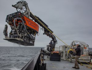 An MBARI remotely operated vehicle being lifted out of the water. The robotic submersible has a bright orange float with the name “Ventana” with MBARI’s gulper eel logo serving as the “V.” Beneath the float is a black metal frame with a silver metal robotic arm on the right-hand side, a plastic tube on the left-hand side, and a black camera in the center. The submersible was photographed during recovery, attached to a black-and-white crane and positioned over the side of a research ship with gray water below and overcast gray sky in the background. A member of the ship’s crew wearing a white hard hat, black sweatshirt, brown pants, and brown boots is standing on the gray deck of a research ship and holding the bright yellow tether connected to the submersible.