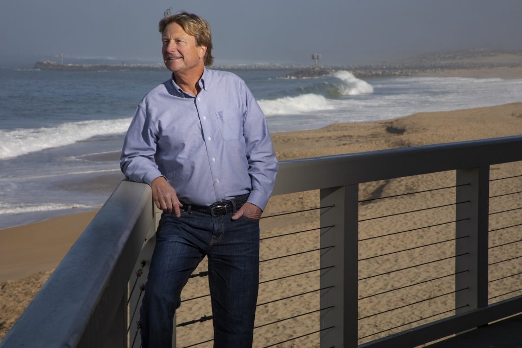 A scientist poses for a photo outdoors, leaning against a metal railing overlooking the beach and ocean.