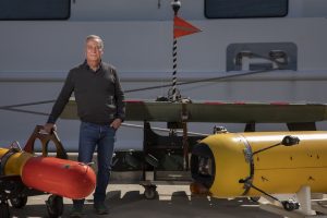 A scientist stands for a portrait outdoors on a sunny day. The scientist has short gray hair and is wearing a gray sweater, blue shirt, blue jeans, and gray shoes. On the left side of the frame is a bright orange underwater robot, and on the right side of the frame is a yellow underwater robot with several cameras. Behind the scientist is a dark green glider robot. In the background is the white metal hull of a research ship.