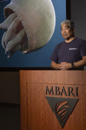 An MBARI researcher stands behind a podium giving a presentation. On the front of the podium is the MBARI logo. In the background are gray walls of a conference room and a projector screen displaying video footage of a large red jelly swimming in open blue water.