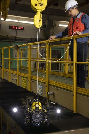 An engineer wearing a hard hat and life vest stands on a yellow fiberglass walkway above a large tank of water, inspecting a portable underwater robot suspended from a crane out of frame.
