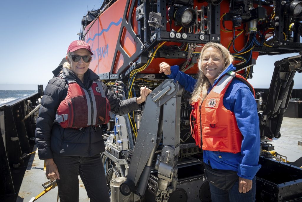 The Monterey Bay Aquarium CEO and the MBARI President and CEO pose for a photo in front of a robotic submersible on the deck of a research ship on a sunny day. Both women are wearing orange life vests.