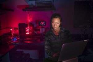 An engineer tests an imaging system in a dark lab. She is standing in front of a computer reviewing data. In the background is engineering equipment emitting red light.