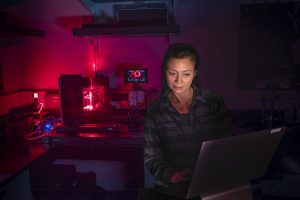 An MBARI engineer with long black hair and a gray-and-black flannel shirt standing in a darkened laboratory observes a computer screen while testing a laser imaging system. In the background is a work bench with a camera and bright red laser projecting into a clear acrylic box filled with water. The room has a dark pink glow.