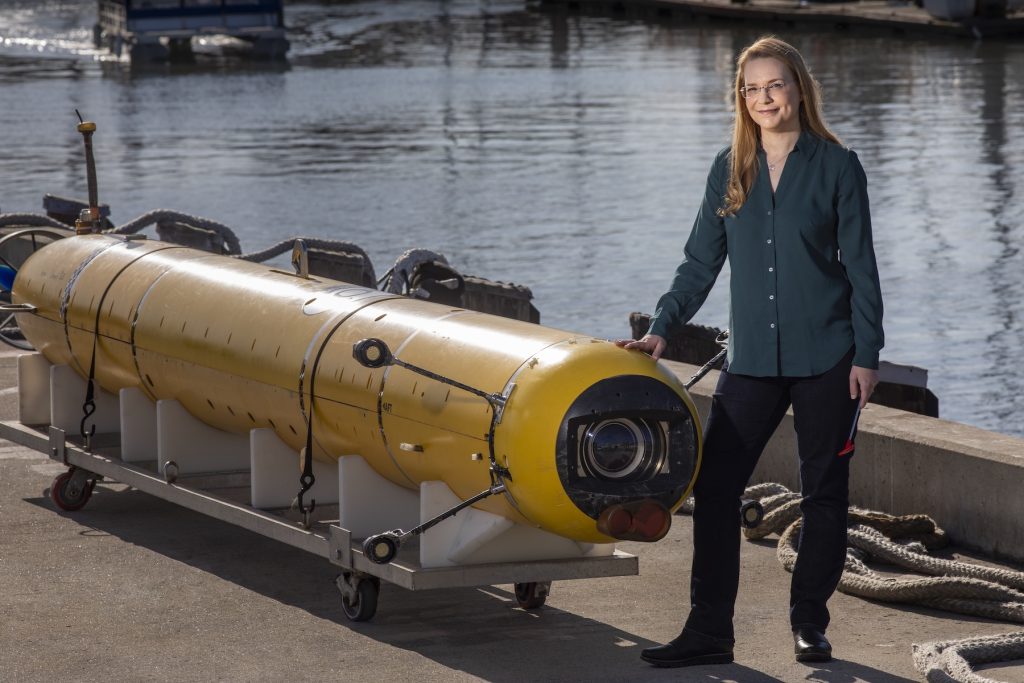 A scientist stands for a portrait photo with an autonomous robot. The scientist has long blonde hair and is wearing glasses, a teal shirt, and dark blue jeans. She is standing next to a cylindrical autonomous robot with a yellow plastic housing, black camera, and four lights sitting on a white plastic cart. In the background are a gray concrete dock and still water in a harbor.