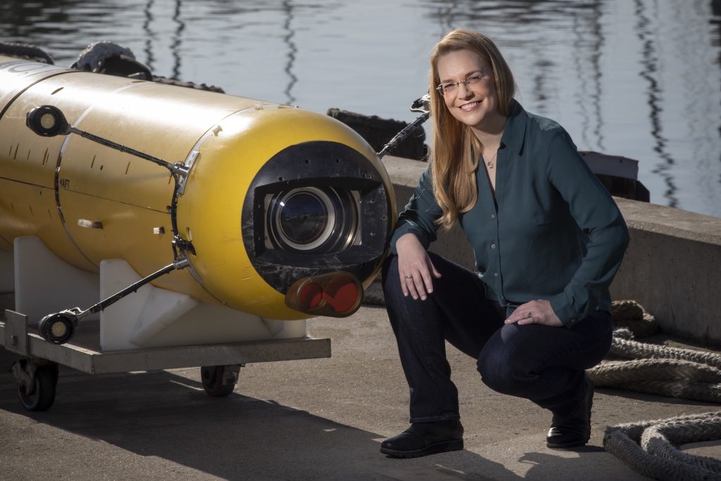 A scientist crouches down for a portrait photo with an autonomous robot. The scientist has long blonde hair and is wearing glasses, a teal shirt, and dark blue jeans. She is standing next to a cylindrical autonomous robot with a yellow plastic housing, black camera, and four lights sitting on a white plastic cart. In the background are a gray concrete dock and still water in a harbor.