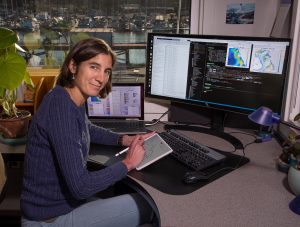 An MBARI researcher with shoulder-length brown hair wearing a navy-blue sweater and blue jeans sits at a desk reviewing oceanographic data. She is holding a white tablet computer with a stylus. On the gray desk are a silver laptop computer and large black monitor displaying multicolored datasets and computer code. In the background is a window overlooking a harbor with ships.
