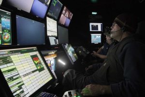 A submersible pilot and aquarium biologist sitting in the dark control room of an MBARI research vessel. The pilot in the foreground is wearing a dark cap, glasses, black vest, navy blue sweatshirt, and dark gray pants, and holding a black joystick. The aquarium biologist in the background is wearing a baseball cap, glasses, a blue vest, and a blue polo shirt. Both individuals are wearing a black headset with earpiece and microphone as they watch a wall of monitors on the left displaying underwater video and data from the submersible.