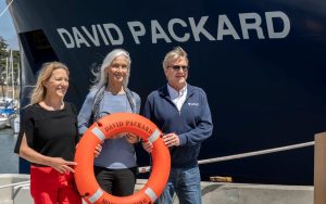 MBARI President and CEO Antje Boetius (left), MBARI Board Chair Julie Packard (center), and outgoing MBARI President and CEO Chris Scholin (right) stand in front of the research vessel David Packard for the ship’s commemoration ceremony. They are holding an orange life ring that reads David Packard and Moss Landing in black capital letters. They are standing outdoors on MBARI’s dock on a sunny afternoon with the blue bow of a research ship with the name David Packard in white capital letters. In the background is Moss Landing harbor.