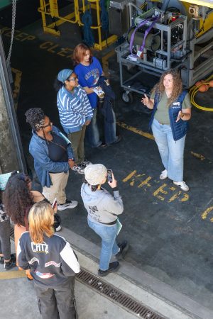 An MBARI researcher speaks with six participants in an ocean career pathways program. She is standing in a loading bay on a gray concrete floor in front of scientific equipment.