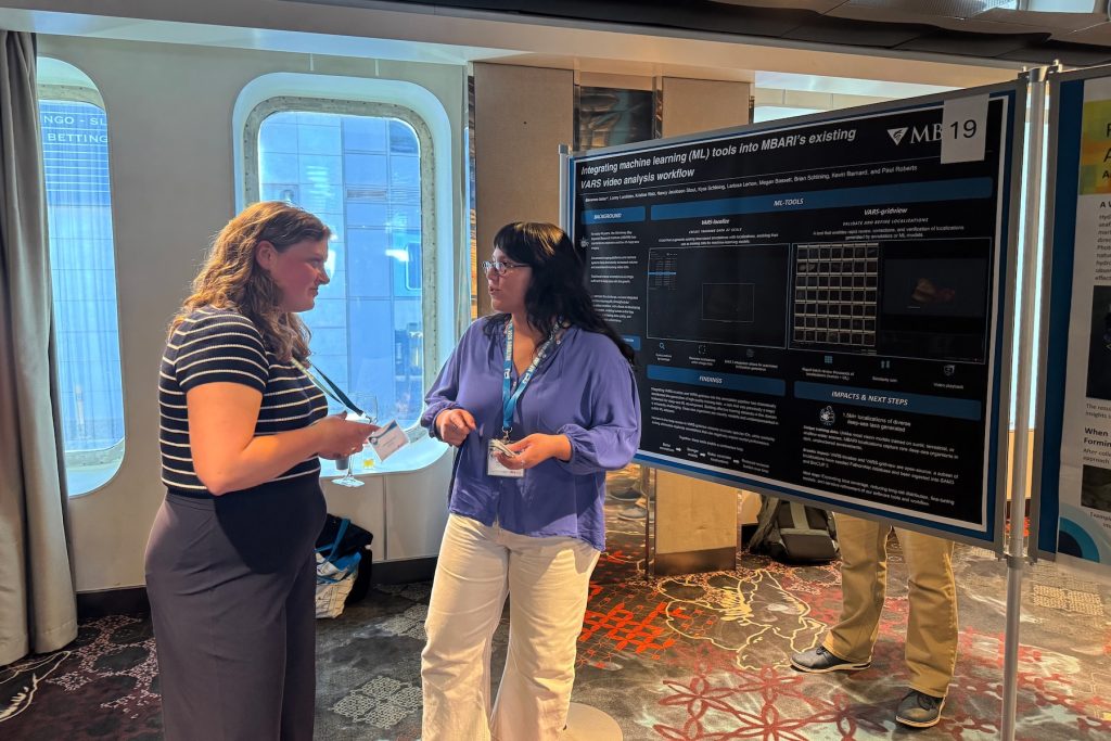 A video researcher presents a poster at a scientific conference. She is standing in front of a black poster with white text mounted on a silver board, and speaking to a conference attendee holding a glass of water. In the background is the interior of a hotel ballroom, with large windows overlooking adjacent buildings.