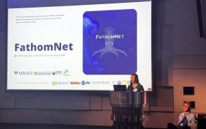 An engineer gives a presentation at a scientific conference. She is standing in front of a rounded silver metal podium. Behind her is a projected presentation slide with a white background, the blue FathomNet Program logo, the names and logos of FathomNet Program members, and black text that reads, FathomNet Accelerating the processing of ocean visual data. In the background are a software engineer seated in a chair and the gray walls of hotel ballroom.