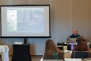 An MBARI engineer wearing a teal fleece jacket and a black t-shirt leads an interactive workshop. The engineer is seated at a table with a silver computer in front of him. In the foreground are workshop participants, seated at tables covered in white tablecloths. In the background is a projector screen displaying a blue software application with a photo of the seafloor on the left and white text annotations on the right. Behind the screen is a gray paneled wall.