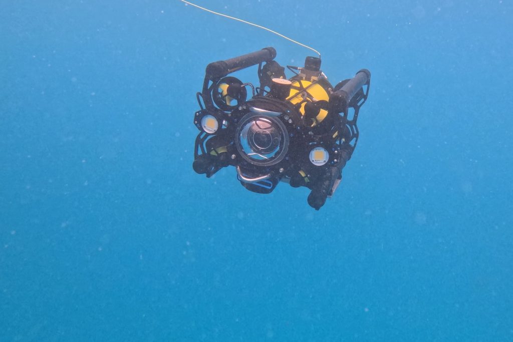 A small portable underwater robot drifts in blue water, tethered by a thin yellow cable to a ship out of frame.