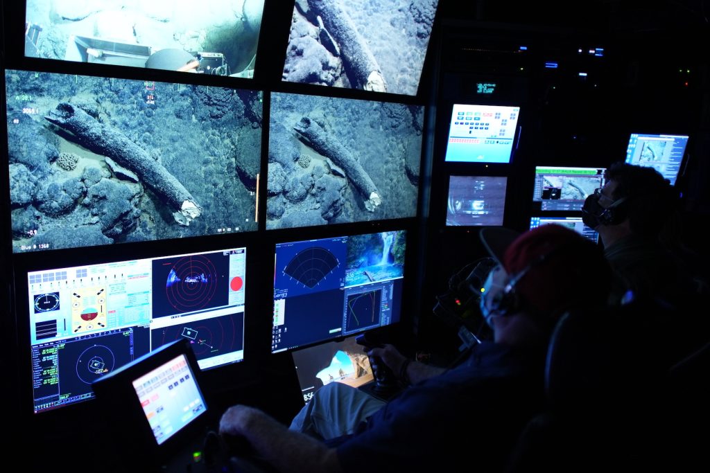 Two MBARI staff in the darkened control room of an MBARI research ship. On the left is an ROV pilot wearing a face mask, a red baseball cap, and headphones and sitting in a chair with a joystick controlling a robotic deep-sea submersible. To the right is a researchers wearing a face mask, glasses, and headphones. Both are watching four television screens displaying the tusk of an ancient mammoth on the deep seafloor. Several other screens display data from the deep-sea submersible.